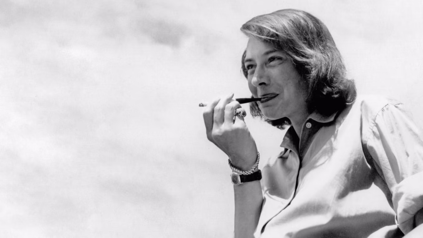 The Price of Salt author Patricia Highsmith smokes a cigarette while smiling in this black-and-white photo taken in the 1960s. She wears a pinky ring on the hand holding her cigarette.
