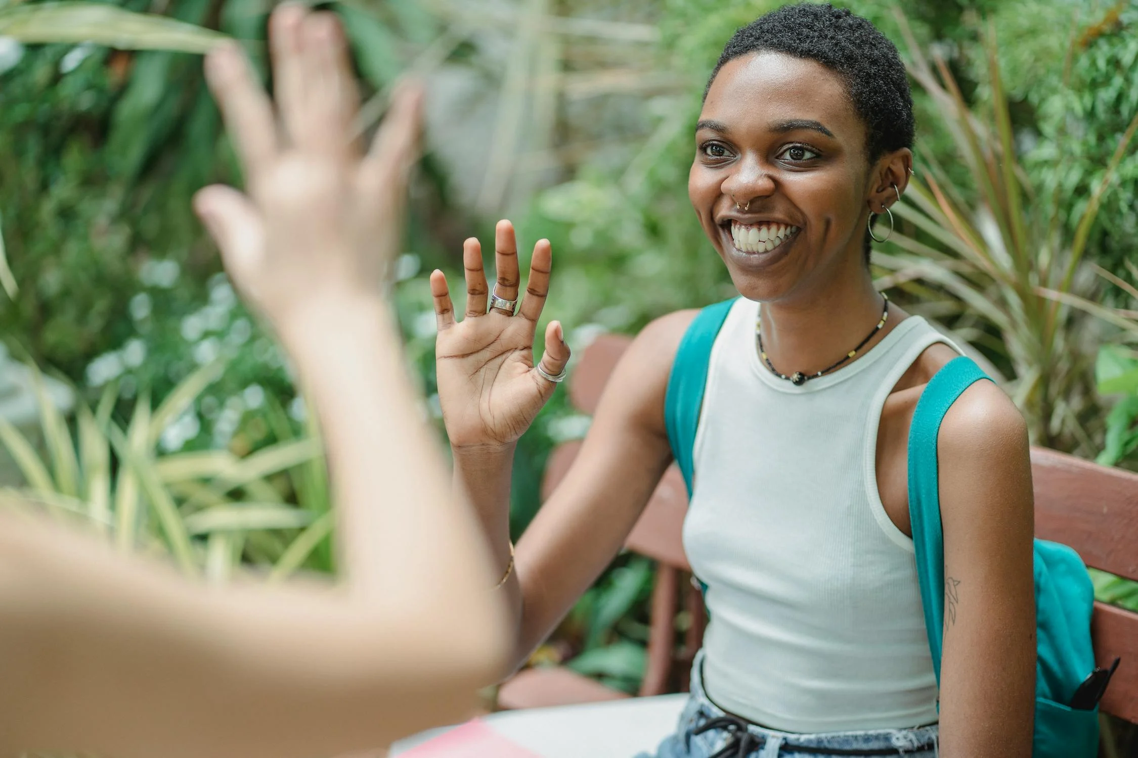Queer woman sitting and waving at a friend. She is wearing many rings and a necklace, reflecting her personal style.