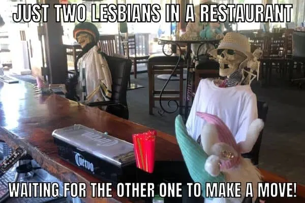 An image of two skeletons sitting far away from each other at a table with meme text reading “JUST TWO LESBIANS IN A RESTAURANT WAITING FOR THE OTHER ONE TO MAKE A MOVE!”