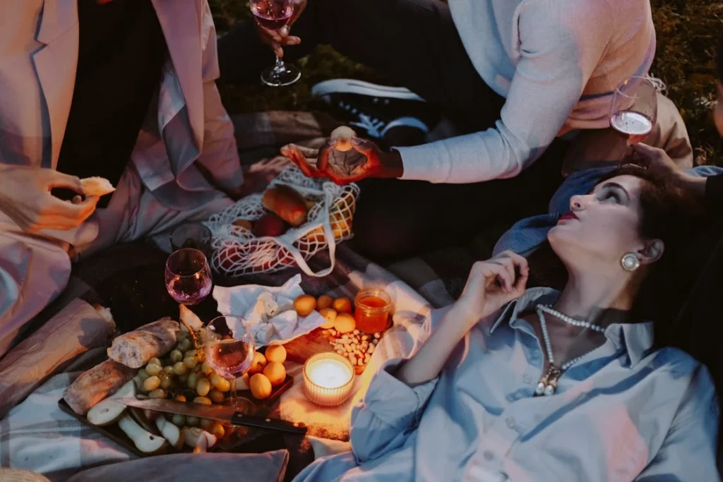 A throuple of people enjoy a cozy outdoor winter picnic with wine, bread, fruits, and candles on a blanket, while one person reclines elegantly, wearing pearls and gazing thoughtfully.