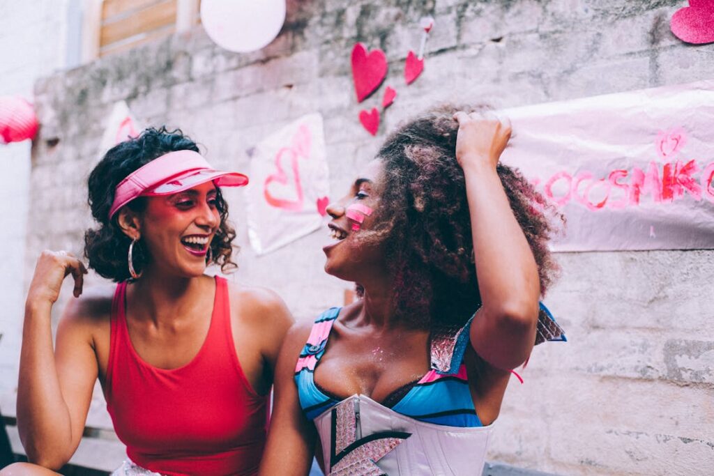 Two friends dressed in vibrant outfits and colorful face paint laugh together against a festive backdrop of pink hearts and decorations, creating a playful and celebratory atmosphere.