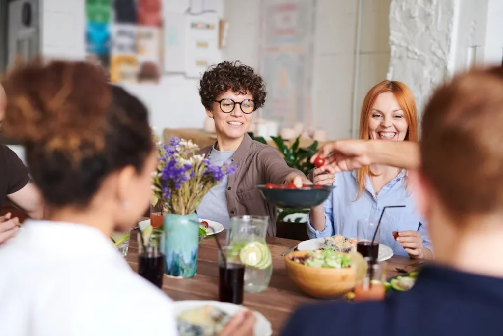 A group of friends gathers around a table, sharing food and laughter, with fresh salads, drinks, and a vase of flowers adding to the warm and inviting atmosphere.