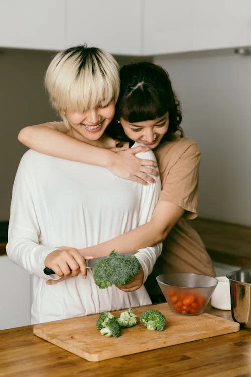 Two women share a joyful moment in a kitchen as one chops broccoli on a wooden board while the other hugs them affectionately from behind, with a bowl of cherry tomatoes nearby.