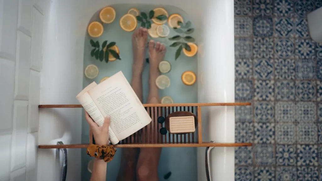 A relaxing bathtub scene features a person soaking with slices of citrus fruits and green leaves floating in the water, while they hold an open book over a wooden bath tray that also holds soap and a candle, set against a patterned tile background.