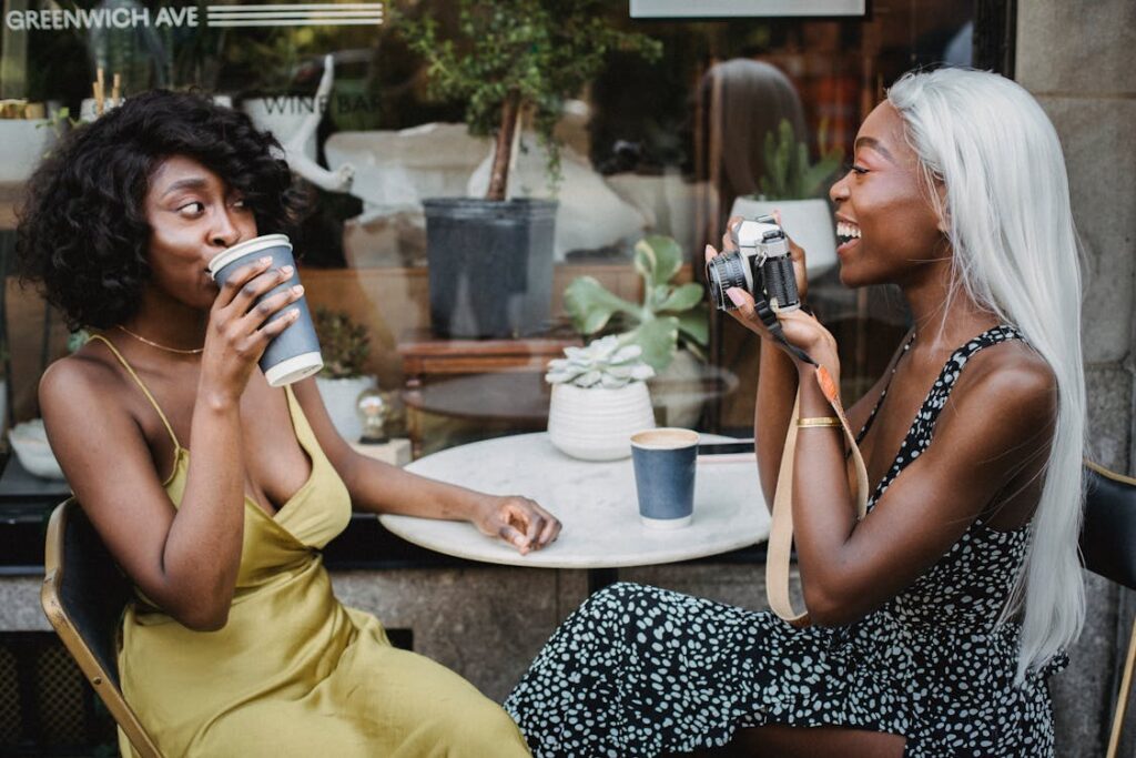Two women enjoy a relaxed moment at an outdoor café, with one sipping coffee and the other playfully holding a camera, both laughing and surrounded by plants on a small table.