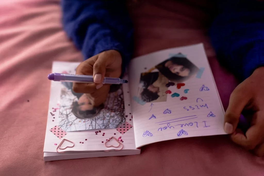 A person wearing a blue sweater writes in a decorated notebook with photos, glitter, and heart designs, featuring handwritten messages like "I love you" and "miss you," resting on a pink surface.